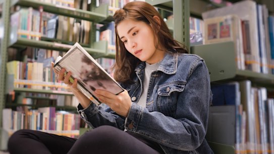 Asian female student sitting on floor in the library, Open and learning textbook from bookshelf in the International College/University Library.