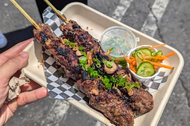 A hand holding a takeaway tray of food with skewers