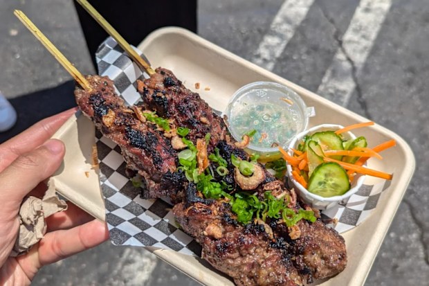 A hand holding a takeaway tray of food with skewers