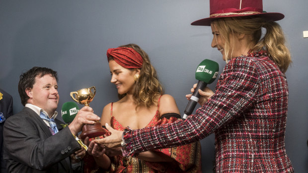 Strapper Stevie Payne gets his replica Strapper's Cup from actor-director Rachael Griffiths, with jockey sister Michelle Payne in the middle on Melbourne Cup Day 2018.