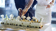 Newlyweds happily cut, laugh and taste the wedding cake. Beautiful Bride and Groom Celebrate Wedding at an Evening Reception Party with Multiethnic Friends. Married Couple Standing at a Dinner Table, Cutting Wedding Cake.