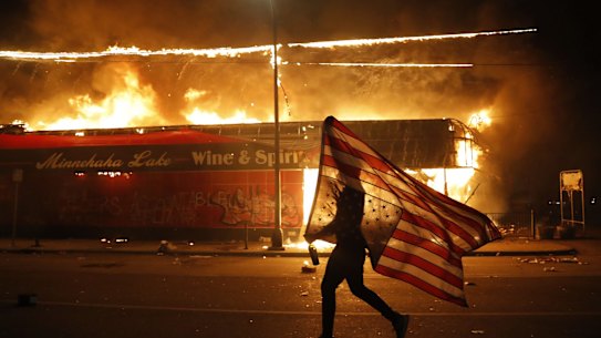 A protester carries the carries a U.S. flag upside, a sign of distress, next to a burning building Thursday, May 28, 2020, in Minneapolis. Protests over the death of George Floyd, a black man who died in police custody Monday, broke out in Minneapolis for a third straight night. (AP Photo/Julio Cortez)