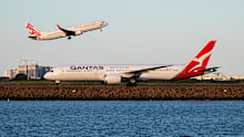 A Qantas Boeing B787-9 plane, registration VH-ZNI, taxiing to the international terminal of Sydney Kingsford Smith Airport after arriving as flight QF28 from Santiago. In the background is a Virgin Australia Boeing B737-8FE plane, registration VH-VUR, taking off as flight VA862 to Melbourne. In the far background are tower blocks. This image was taken from Kyeemagh, Botany Bay on a sunny afternoon approaching sunset on 11 April 2025.