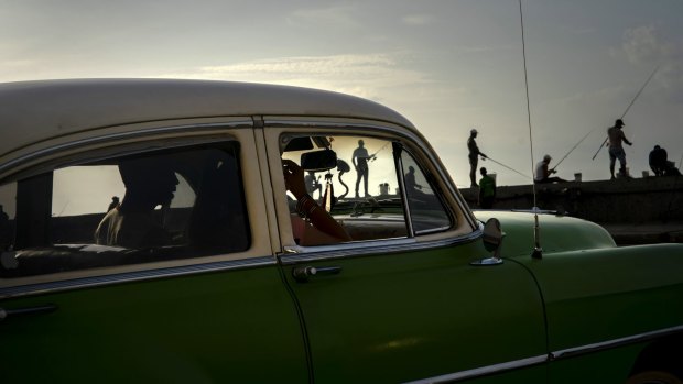 A couple in a classic American car drive along the Malecon seawall past fishermen at sunset in Havana, Cuba.