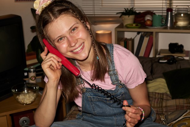 Girl smiling and talking on red phone while sitting cross-legged in cozy room filled with various knick-knacks and colorful posters hanging on wall, creating a retro atmosphere