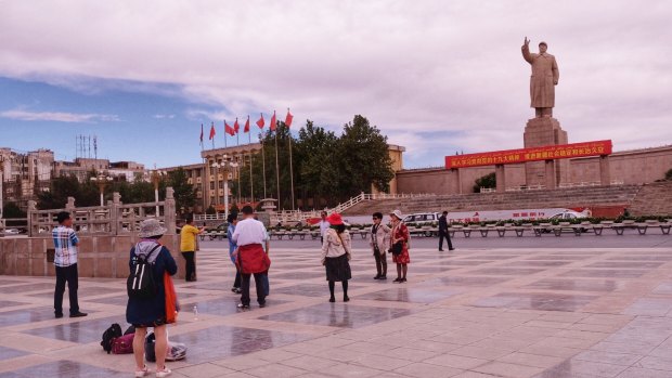 Han Chinese tourists take photos of a monumental sculpture of Chairman Mao Zedong. A big red sign under the statue encourages people to "implement the spirit" of the communist party and promote "social stability" in Xinjiang.