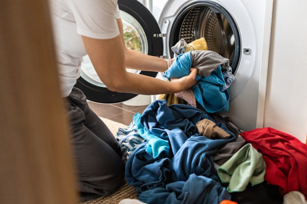 A woman places colourful dirty clothes into a front loader washing machine.