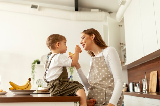 young mother gives cookies to her little son in the kitchen, 2-year-old boy eats with his parent, woman in apron feeds the child