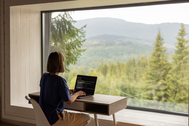 Woman works on laptop while sitting by the table in front of panoramic window with great view on mountains