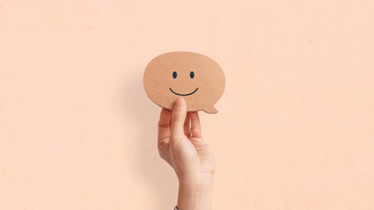 Hand holding happy face smiling comment in speech bubble on pastel background. Happiness. Generic. 