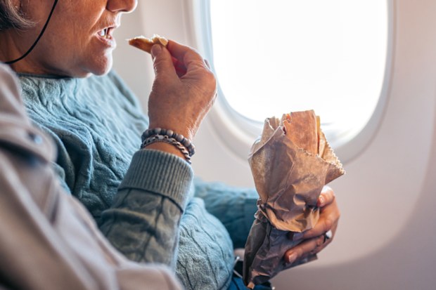 Senior woman eating a sandwich while riding in an Airplane. Her senior husband is beside her.