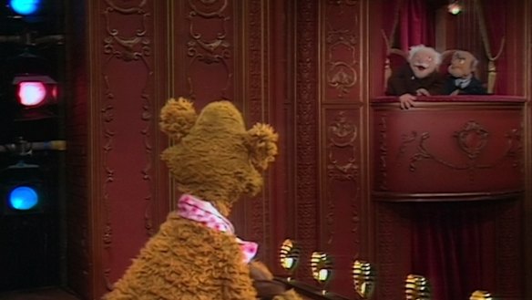 Statler and Waldorf heckle Fozzie Bear on stage in The Muppet Show.