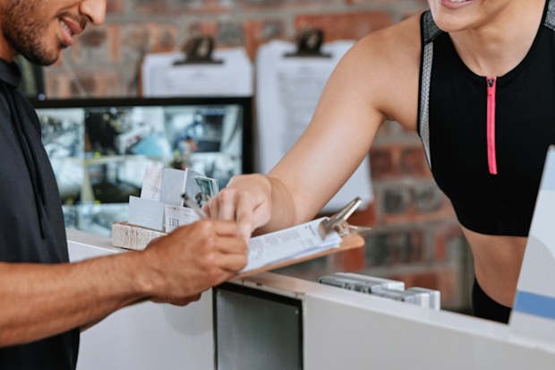 Asian man signing up for gym membership with caucasian trainer. Young coach behind reception pointing and showing where to sign. Two active and fit people standing together in health and fitness club