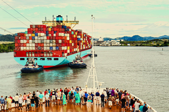 Tug boats help a container ship enter the canal, watched by passengers on a cruise ship. 