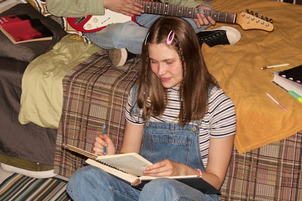 Two girls sitting on carpeted floor and bed in well-decorated retro bedroom, one studying while the other playing guitar, creating a relaxed and productive atmosphere