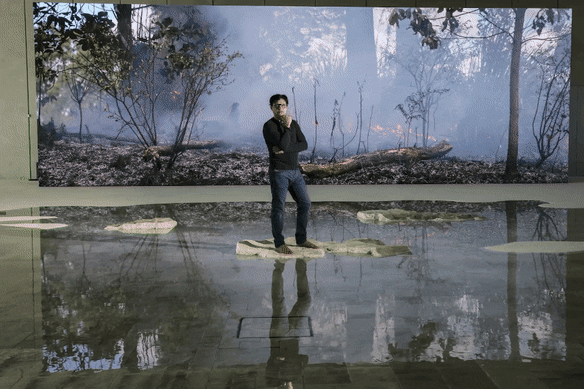 Ivan Sen in front of the LCD screen showing images from the exhibition Overflow at Parramatta Powerhouse. 