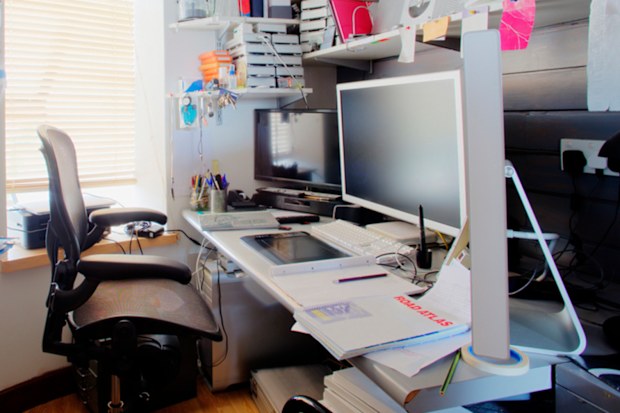 A shot of a messy desk in a home office, the room is small and cluttered.