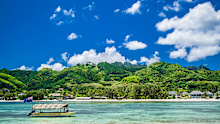 Rarotonga coast during a sunny day. One boat with Cook Islands flag on the sea. Blue sky with clouds and turquoise water. Partly cloudy. Palm trees and resorts in distance.