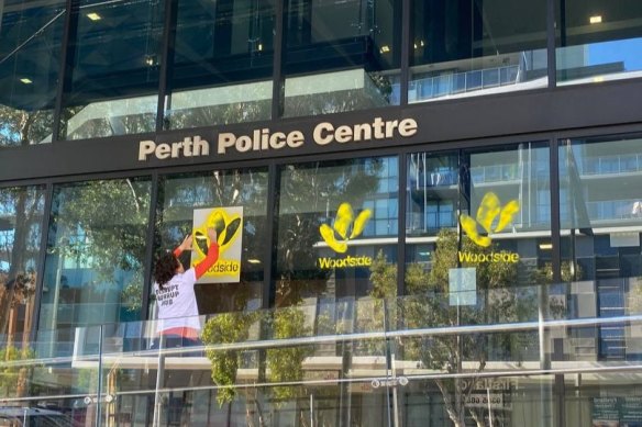 Violet Coco sprays the Woodside logo in yellow paint on the front of the Perth Police Centre in protest at the escalating police crackdown on climate protest in WA.