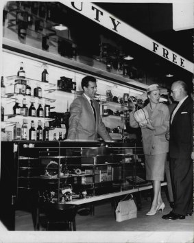 A passenger makes a purchase at the Mascot Duty Free store in 1961. The good old days of saving a lot on duty free purchases are gone.
Salesman Mr. Gerry Boag (left) shows Miss Margot Lee and London-bound passenger Mr. T. Renenson a bottle of wine in the new duty-free shop at Kingsford Smith Airport which opens today. Duty-free goods are available only to bona fide travellers leaving Australia. Prices range from 25 per cent more than 50 per cent below the retail figures in other stores where duty must be paid. May 2, 1961. 