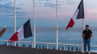A man poses for a picture next to French flags flying at half mast on the Promenade des Anglais in Nice, France.