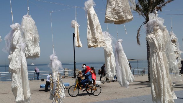 In this April 22, 2017 photo, a veiled woman with her daughter, center, rides a bicycle past white wedding dresses displayed by the Lebanese NGO Abaad, to protest against article 522 in the Lebanese penal code that stipulates that a rapist is absolved of his crime if he marries his victim, on the Beirut corniche, Lebanon. 