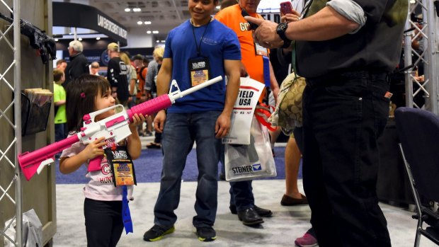 A young girl, who is already an NRA lifetime member, gets her photo taken holding a rifle painted pink and white in the trade booths at the convention.