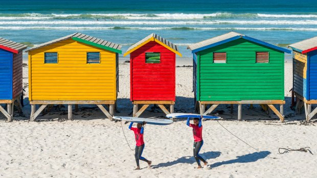 Smiling surfers walking by the beach cabins and huts of Muizenberg, a famous surfing spot near Cape Town.