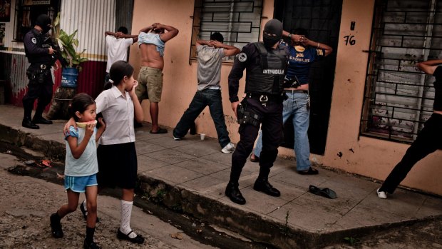 School children walk by as heavily armed members of a police anti-gang force search men in El Salvador.
