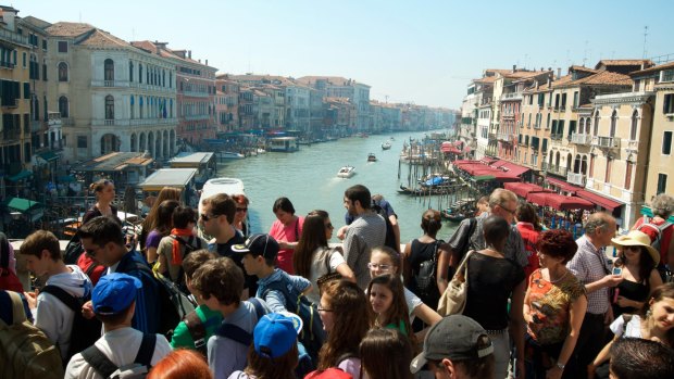 Crowds of tourists pass along the Rialto Bridge against a view of the Grand Canal.
