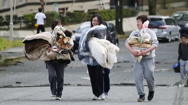 College students carry blankets and foods toward a  shelter as a damaged apartment building is seen in the background after an earthquake in Minamiaso village on Saturday. 