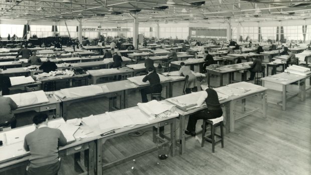 Draftsmen bending over desks in a large warehouse in Fishermans Bend.
A banner at the front of the large room provides cheerful motivation: "Victory begins on the drawing board!" it reads.