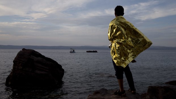 A refugee wrapped in a thermal blanket looks as a dinghy full of migrants approaches the coast of Lesbos.