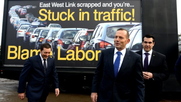 Prime Minister Tony Abbott, flanked by Victorian Opposition Leader Matthew Guy, left, and local member Michael Sukkar, stand in front of a "Blame Labor" sign.