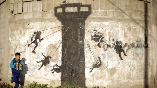 A Palestinian child walks past a mural of children using an Israeli army watch tower as a swing, said to have been painted by British street artist Banksy, on the remains of a house destroyed during Operation Protective Edge in the northern Gaza Strip town of Beit Hanoun.
