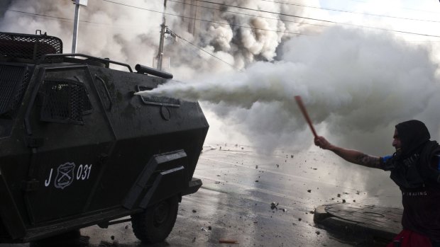 A hooded protester waves a stick at an anti-riot vehicle spraying bursts of tear gas, in front of a burning building near Congress, where President Michelle Bachelet was presenting her state-of-the-nation report.