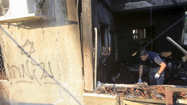 An Israeli police officer inspects the charred remains of the house, with graffiti clearly visible.