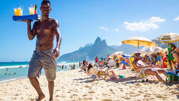 A beach vendor selling caipirinhas calls out to  customers on Ipanema Beach with Two Brothers mountain backdrop.