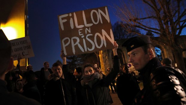 A demonstrator holds a board reading: Fillon in jail, as Francois Fillon delivers a speech during a campaign meeting.