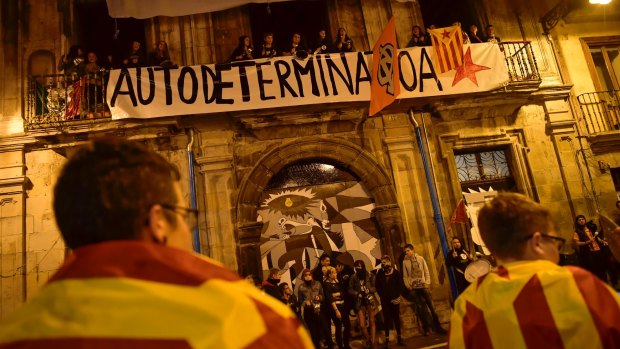 Pro-independence supporters wrapped in ''esteleda'' or Catalan flags stand in front a balcony while people protest with a banner reading ''self determination''.