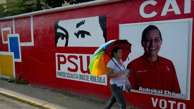A pedestrian walks past a mural depicting the eyes of the late Venezuelan President Hugo Chavez, and an election poster promoting his cousin Asdrubal Chavez, a United Socialist Party of Venezuela congressional candidate, in Sabaneta.