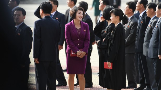 A young woman wearing a modern, brightly-coloured outfit and red clutch purse waits next to others in traditional Korean dresses in Pyongyang. Designer styles, in colours, and expensive jewellery are on the rise in the city. 