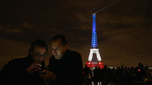 Parisians look at a mobile phone with the Eiffel Tower illuminated in red, white and blue in the background.