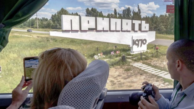 A tour bus, operated by Chornobyl-Tour, drives past a Soviet-era sign marking the entrance to the abandoned city of Pripyat.