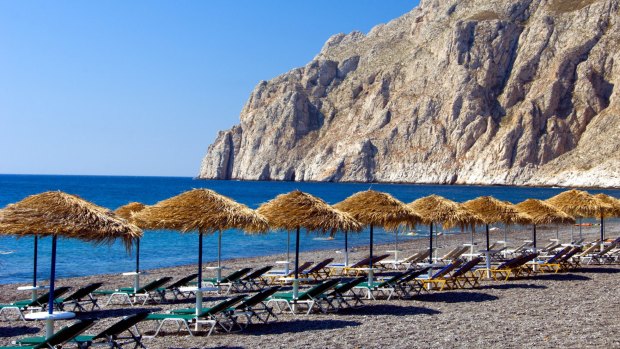 The black sand beaches with umbrellas and lounge chairs in Kamari on the Greek Island of Santorini.
