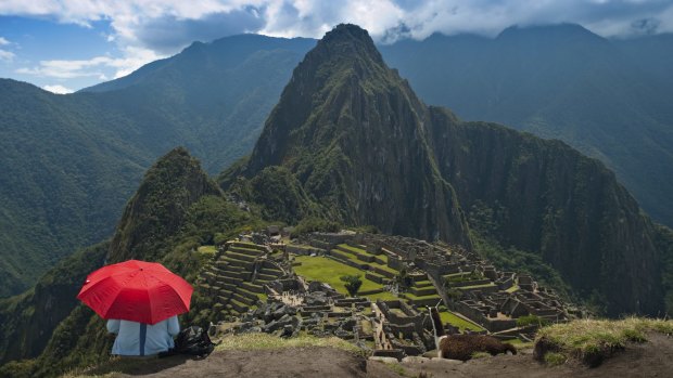 A tourist under the shade of a red umbrella looks at Machu Picchu.