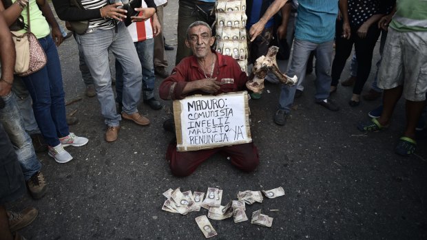 A man holds a cow bone and sits next to a sign that reads "Maduro: unhappy communist quit now", while people throw 100-bolivar banknotes on the ground in El Pinal.