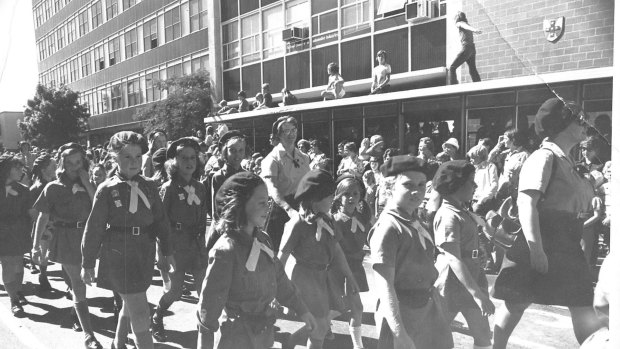 A parade of Guides and Brownies in Canberra in 1964. Note the casual OH and S vibe at the time as kids scramble on to rooftops to watch the passing parade. 