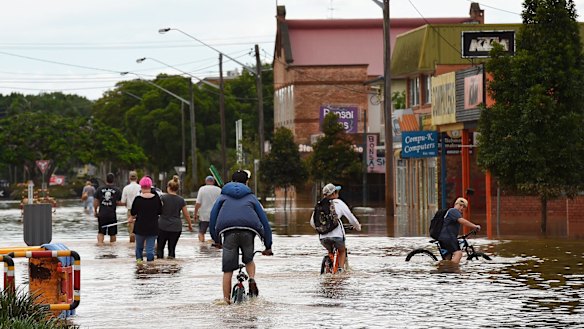 Teenagers cycle behind a group of people with brooms wading through the water of the flooded streets of Lismore.