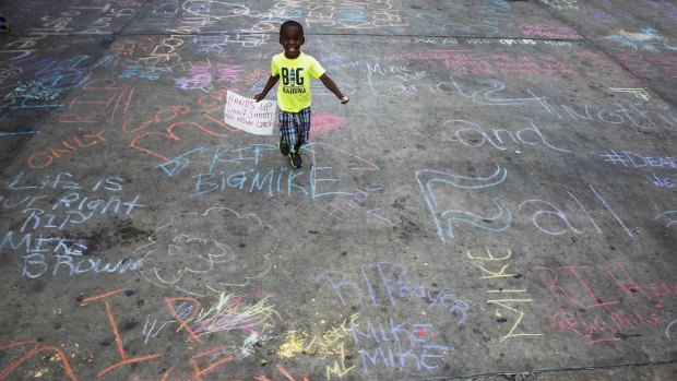 A young boy carries the slogan "hands up, don't shoot, no more lies" on a sign, as he runs through a car park emblazoned with messages of remembrance related to the shooting of Michael Brown.
